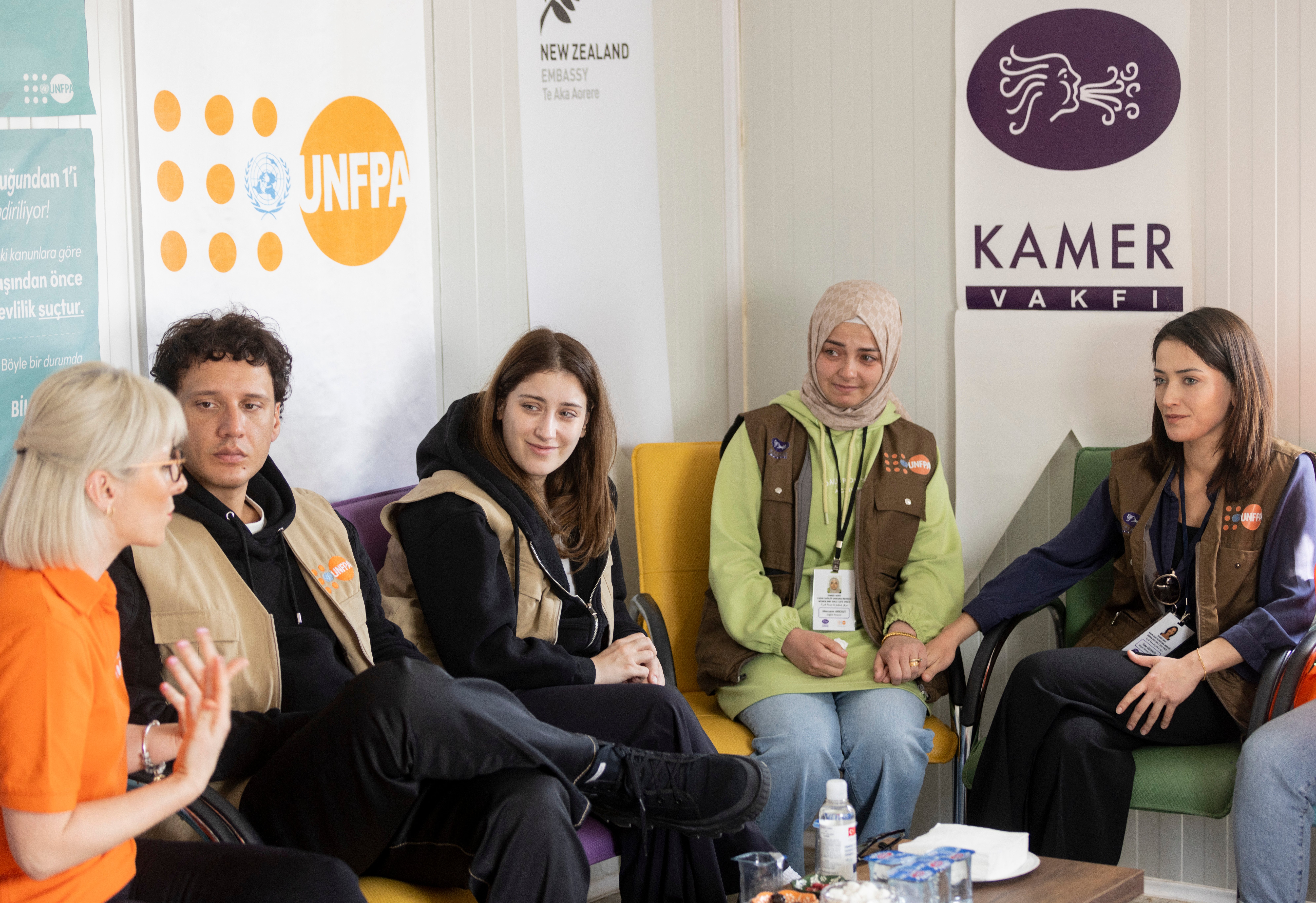 UNFPA Türkiye İyi Niyet Elçileri Hazal Kaya ve Edis, Kahramanmaraş'taki Kadın Sağlığı Danışma Merkezi’ni ziyaret ederek yararlanıcılarla görüşmektedir. Arka duvarda ise UNFPA, Yeni Zelanda Büyükelçiliği ve Kamer logolarının yer aldığı bayraklar yer görünür.