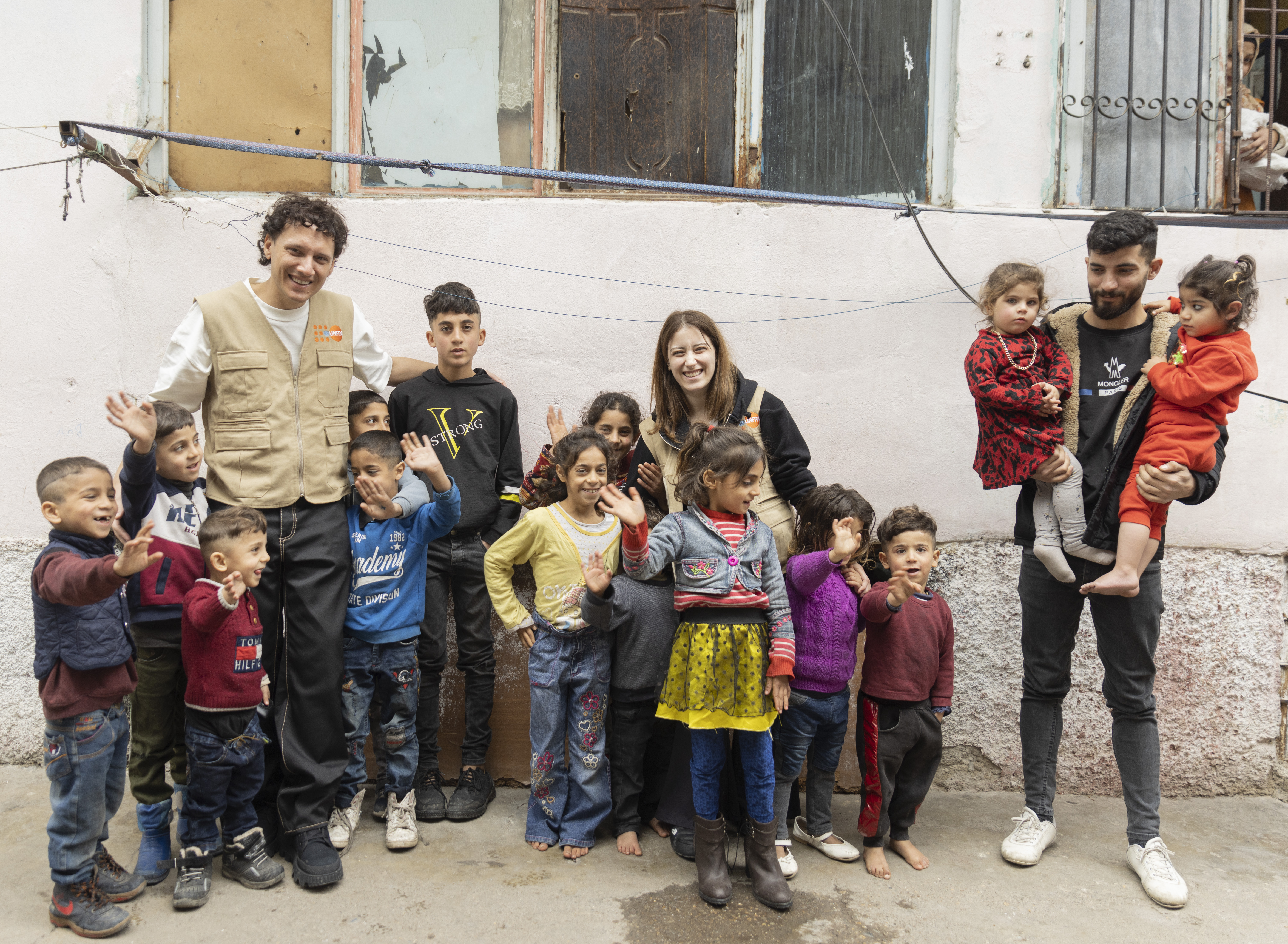 UNFPA Türkiye İyi niyet elçileri Hazal Kaya ve Edis, Kahramanmaraş’taki depremzede aileleri ziyaret ediyor, kalabalık bir çocuk grubuyla birlikte açık alanda poz veriyorlar. 
