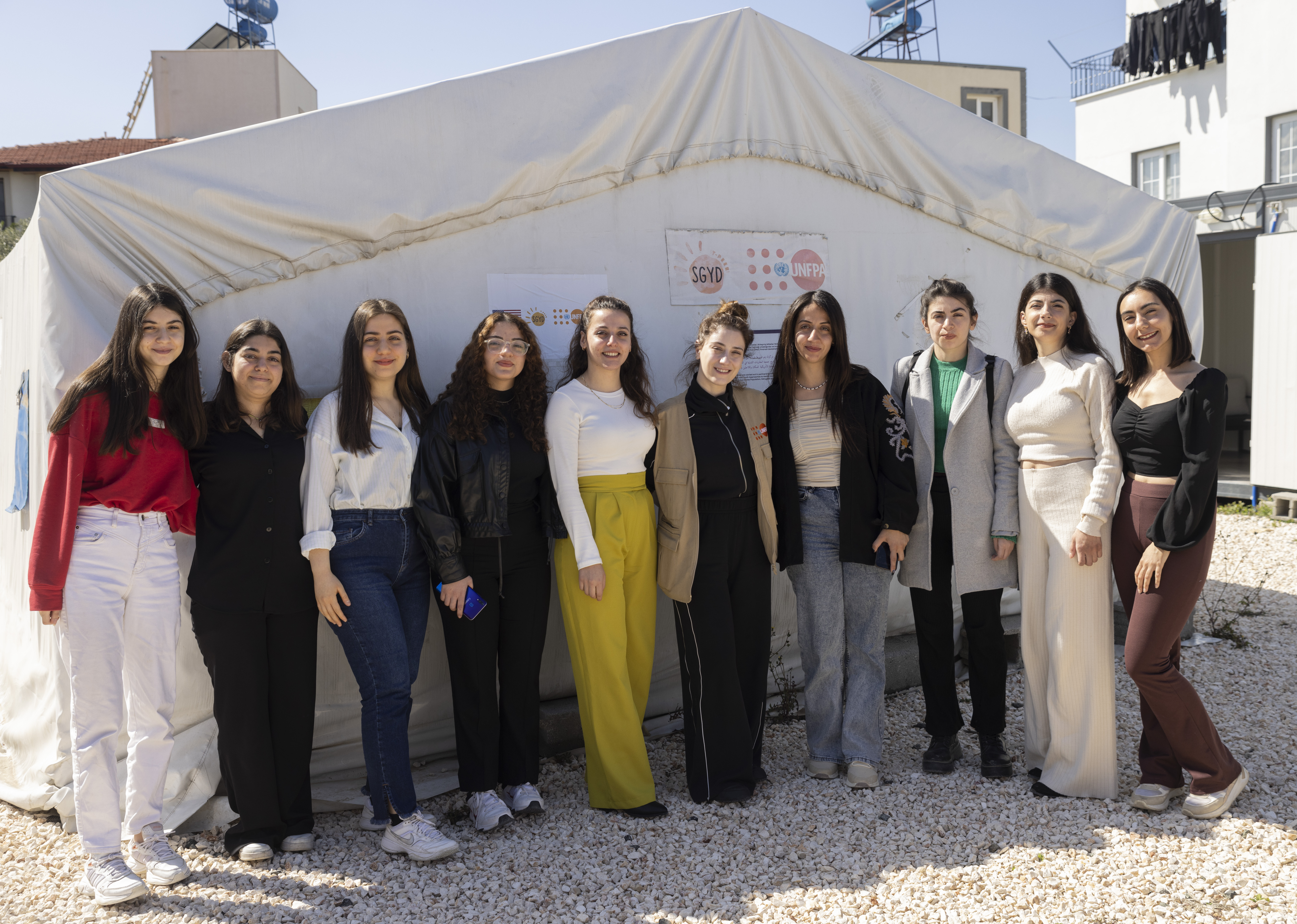 UNFPA Türkiye İyi niyet elçisi Hazal Kaya, bir grup genç kadınla birlikte Hatay Samandağ’daki Gençlik Merkezi önünde poz vermektedir. Arkalarında UNFPA, SGYD ve State PRM logosu görünmektedir.