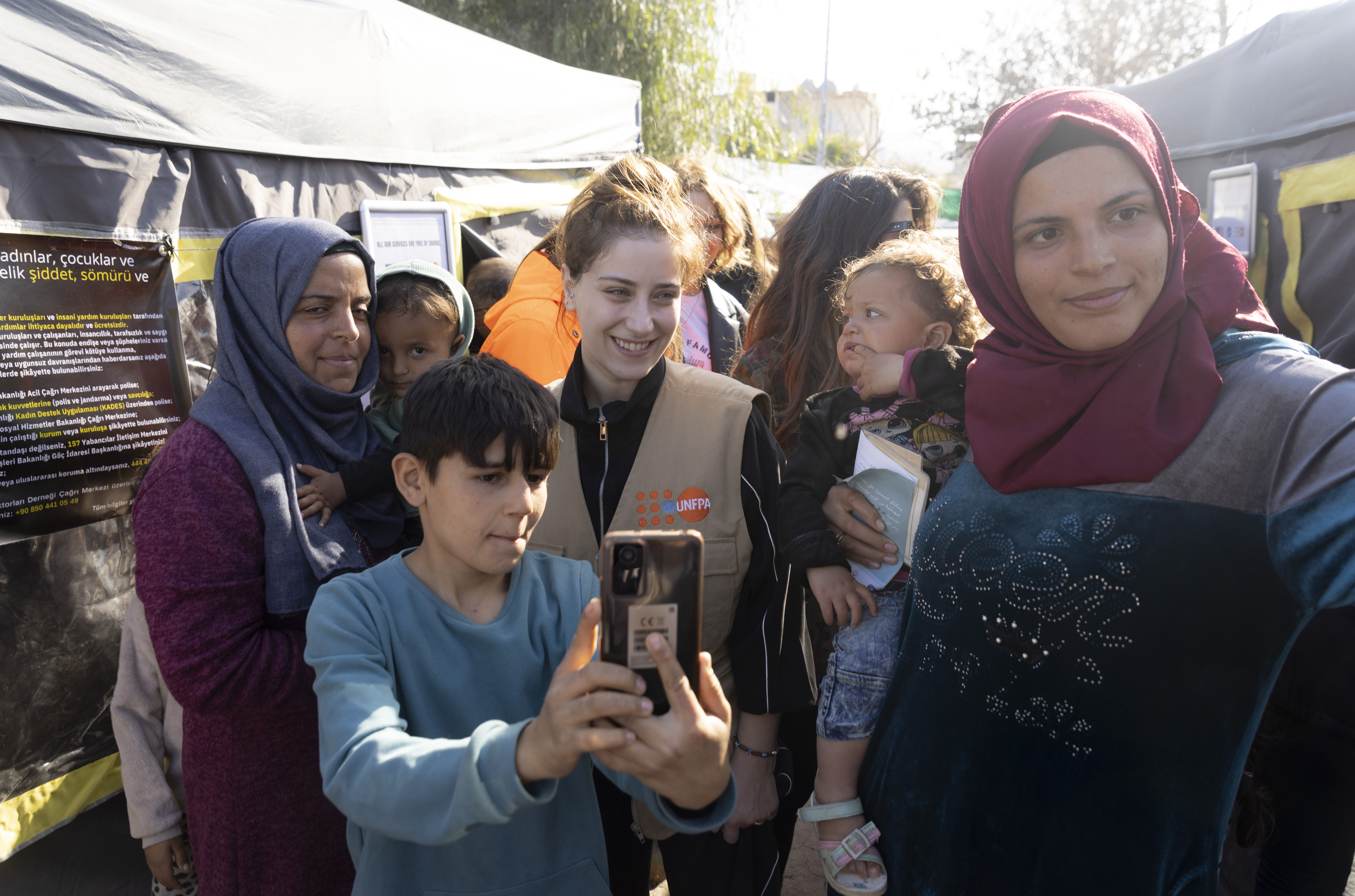 UNFPA Türkiye İyi niyet elçisi Hazal Kaya, bir grup kadın ve çocukla birlikte çadırların önünde poz vermektedir. 