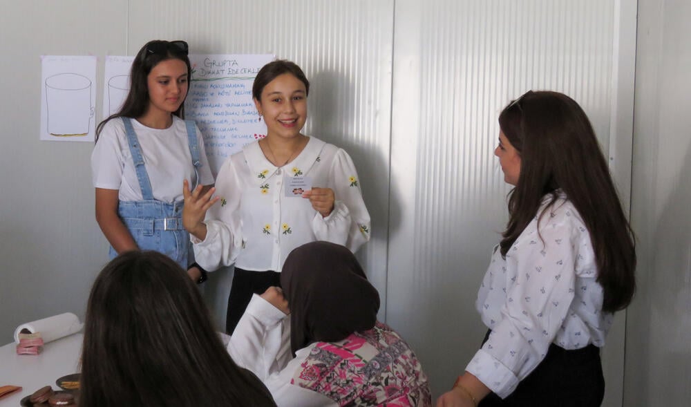 The photo depicts three girls standing and to sitting down inside a classroom. The girl standing in the middle is holding a card and playing a game with her classmates.