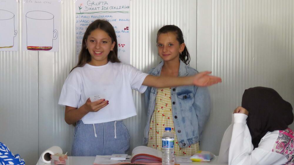 Two girls are standing and making a gesture inside the classroom while playing a game with their classmates.