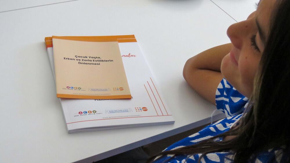 A girl is sitting on a desk, which has a book and a brochure about prevention of early, forced and child marriages.