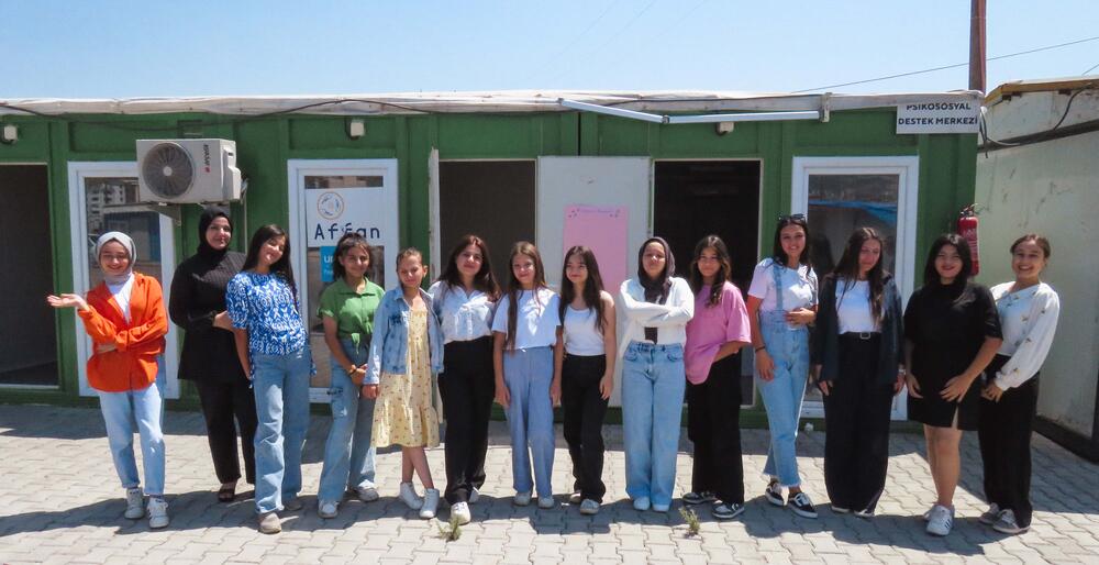 Thirteen girls and their trainer is in front of a container office, posing to camera.