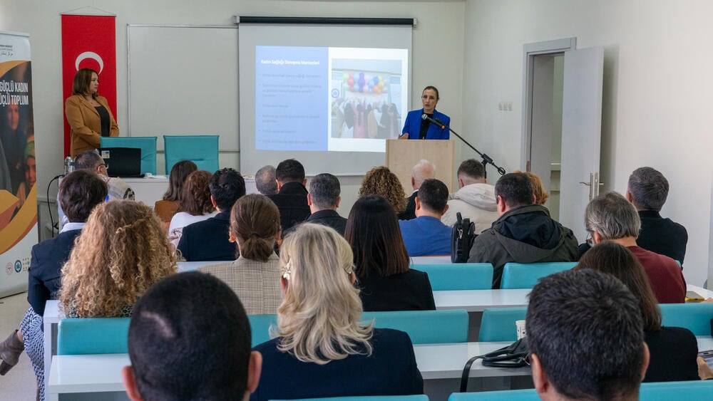 Bir kadının konferans odasında konuşma yaptığı görülüyor. İnsanlar oturmuş ve kadının sunum konuşmasını dinliyorlar.