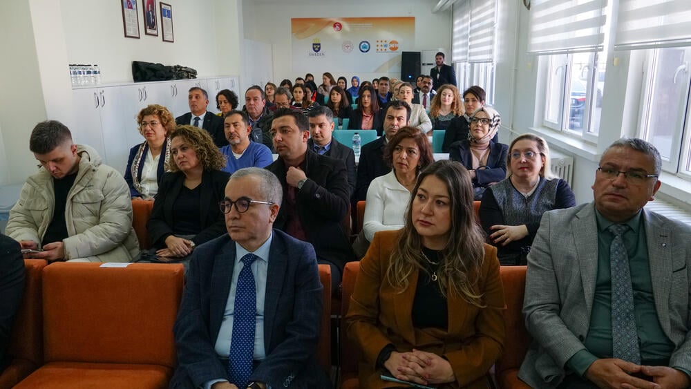 İnsanların oturduğu ve önlerindeki sunumu dinlediği bir oda, insanlarla dolu olarak görülmektedir.