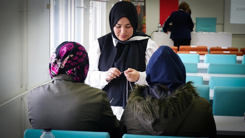 Three women in a room are in the focus. One facing the camera is knitting, and the other two are watching her. 