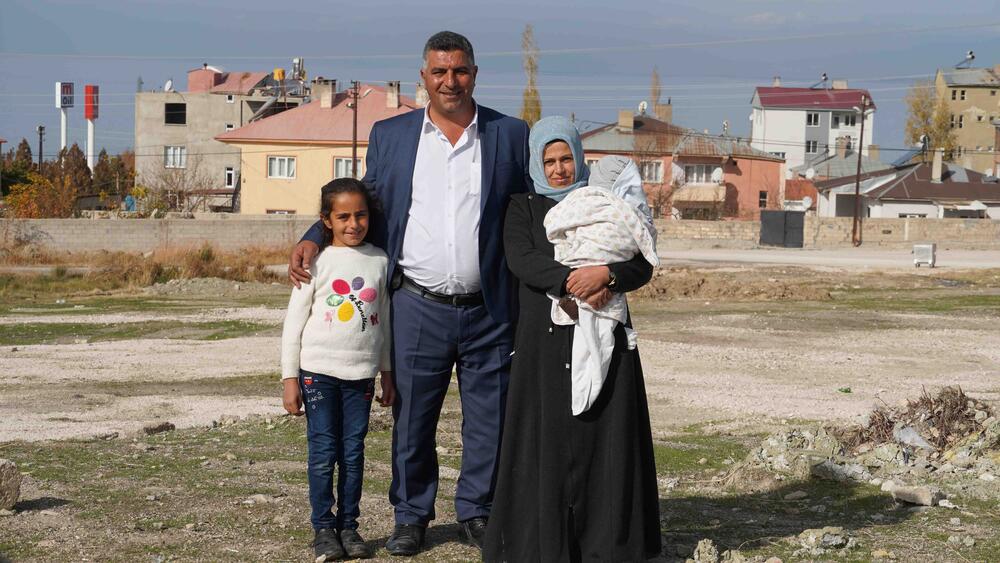 A family of four comprised of a mother, a child, a baby, and a father are standing on a field with a rural settlement in the background.