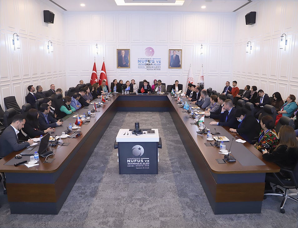 Geniş açılı bir çekimde, iyi aydınlatılmış bir konferans salonunda U şeklinde bir masa etrafında oturan çok sayıda katılımcıyla resmi bir toplantı görülmektedir.