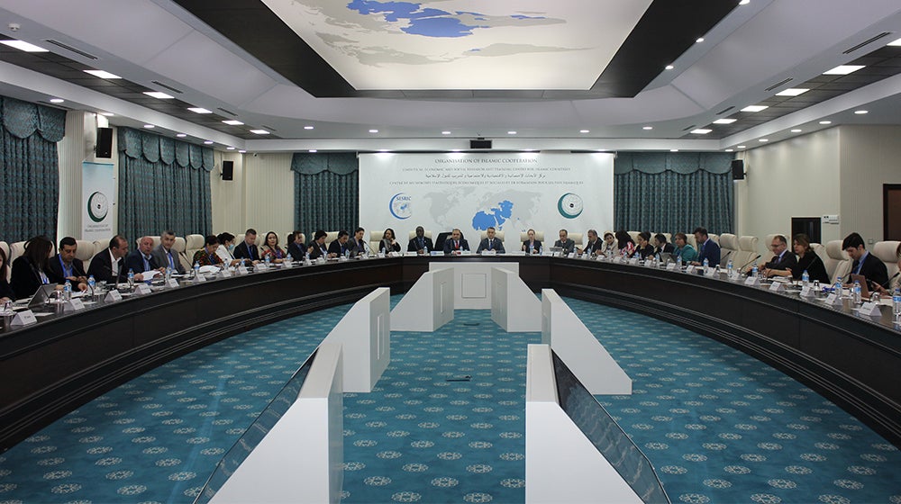 Geniş açılı bir çekimde, büyük, kavisli bir masa etrafında oturan ve bir sunum ekranına bakan çok sayıda katılımcıyla resmi bir konferans salonu görülmektedir.