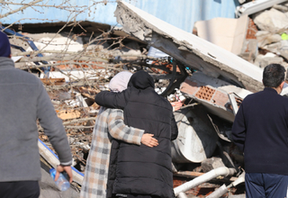 Two women hugging in sorrow in front of a collapsed building