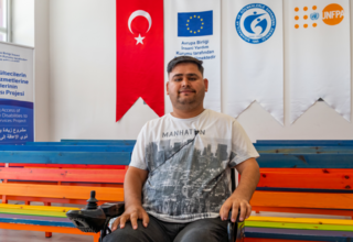 Mohammed Edris sitting in front of a colorful bench, Turkish Flag, EU, ASAM and UNFPA is seen behind.