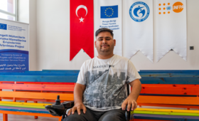 Renkli bir bankın önünde oturan Muhammed Edris'in arkasında Türk Bayrağı, AB, ASAM ve UNFPA logoları görülüyor.