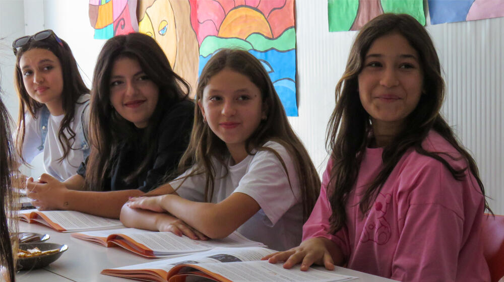 The image depicts four girls sitting inside a classroom while smiling.