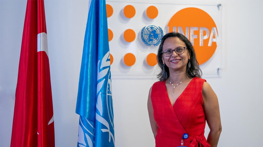 UNFPA Turkiye Representative Mariam Khan is standing in front of a wall that has the logo of UNFPA. Next to the woman are two flag, one of Turkiye and the other United Nations