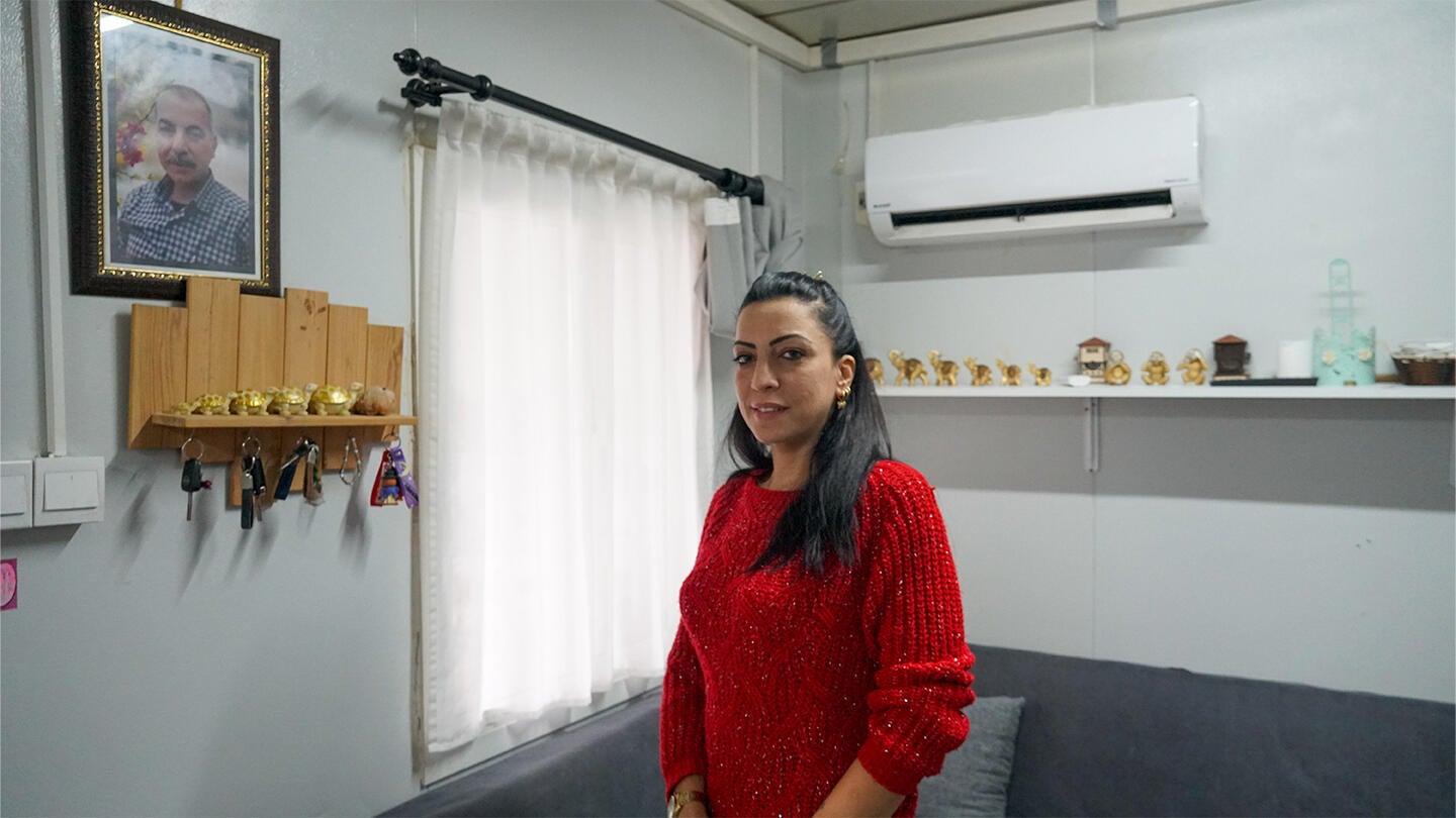  A woman in the center is standing still with a smile. The background shows the room inside a container house.