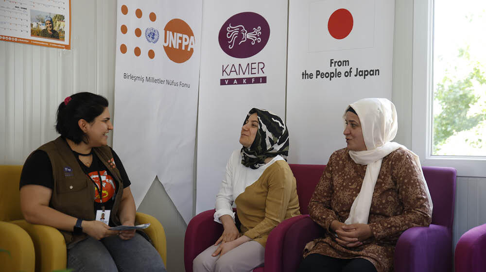 In the center, three women, one wearing a UNFPA jacket, are talking. Behind them UNFPA, KAMER, Government of Japan logos seen