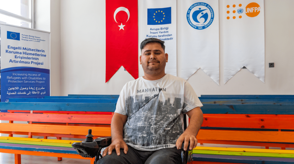 Mohammed Edris sitting in front of a colorful bench, Turkish Flag, EU, ASAM and UNFPA is seen behind.